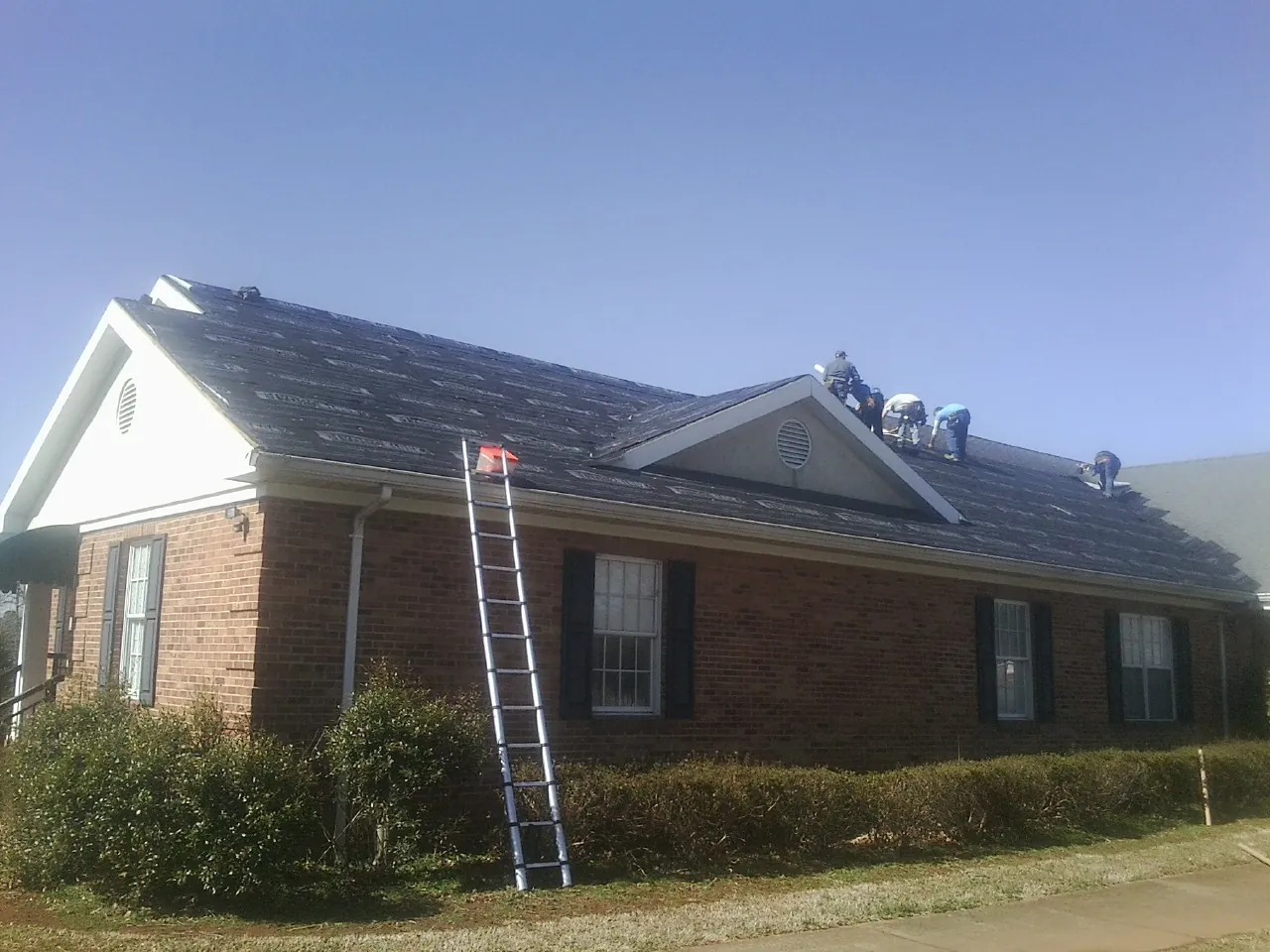 Crew tearing shingles off a brick ranch roof with a ladder set — Metal Roof Repair work by our crew in Peru